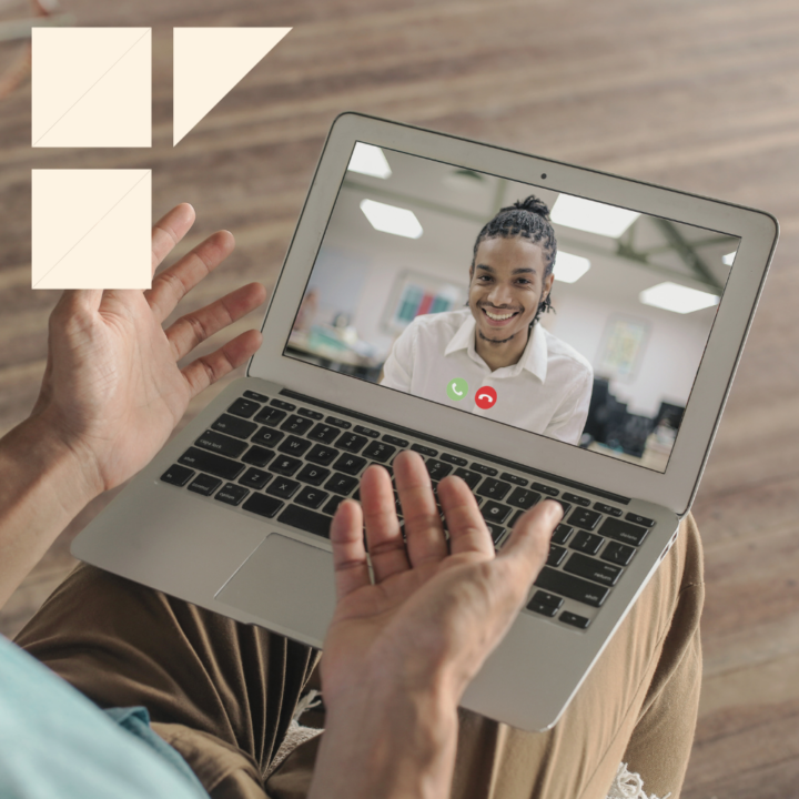A person participating in a virtual interview on a laptop, engaging in a video call with a smiling candidate wearing a white shirt in a well-lit office setting. The interviewer's hands are visible, gesturing during the conversation.