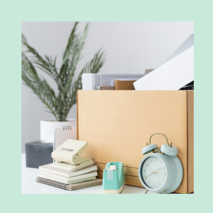 A cardboard box filled with office supplies, including folders, notebooks, and a stapler, sits on a desk. A vintage-style alarm clock and other desk essentials are placed nearby, symbolizing someone packing up their belongings, possibly after resigning from a job.