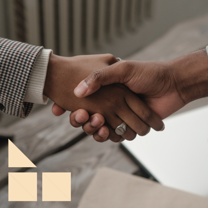 A close-up of two people shaking hands, symbolizing agreement or partnership. One person is wearing a checkered blazer with a white cuff, while the other has a silver ring on their finger. A laptop and documents are visible in the blurred background, suggesting a professional or business setting.