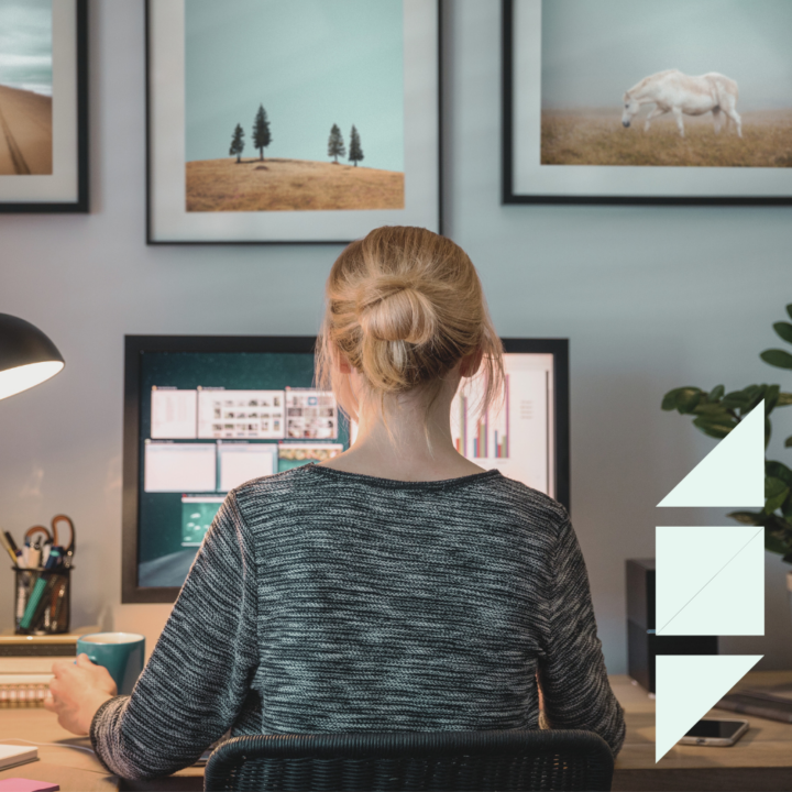 A woman sits at a desk in a home office, working on a computer with multiple screens open. She holds a coffee mug while focusing on her work, suggesting she is starting a new remote job. The workspace is decorated with framed nature prints, plants, and office supplies, creating a productive and cozy environment.