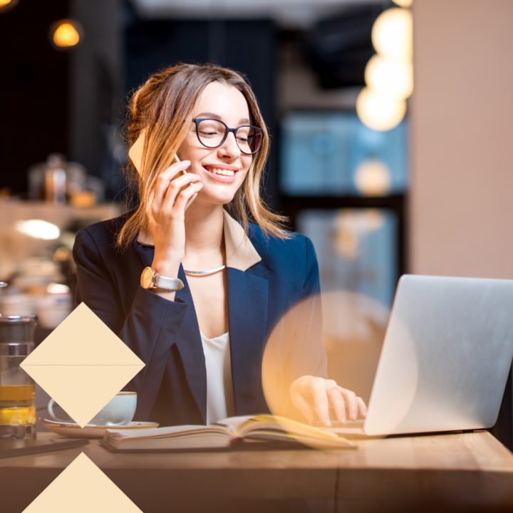 A young professional woman wearing glasses and a navy blazer sits at a café, talking on the phone while working on her laptop. She is smiling and has an open notebook in front of her, suggesting she is engaged in career planning or a job search.