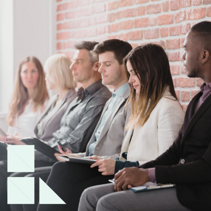 A group of professionally dressed candidates sits in a row against a brick wall, waiting for their job interviews. Some are reviewing notes or using tablets, while others appear focused and prepared. The setting suggests a competitive hiring process.