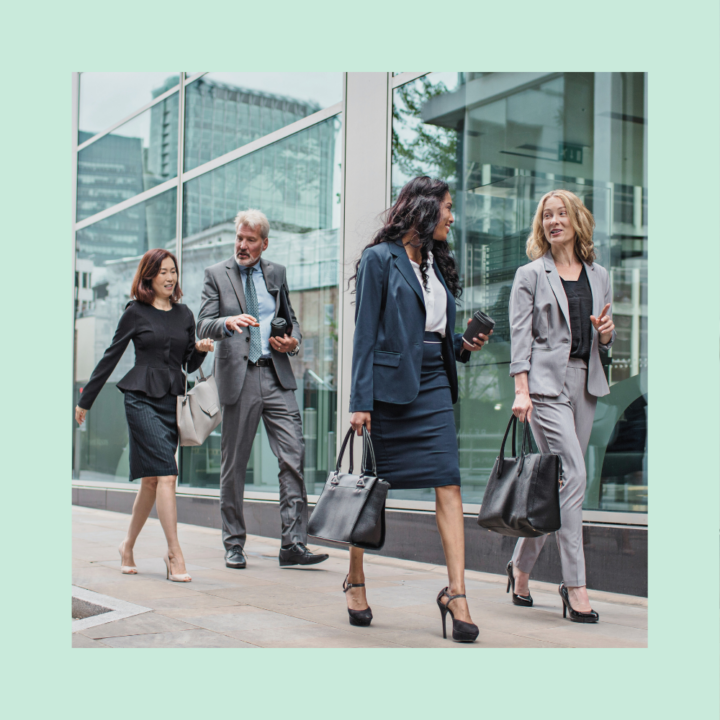 A group of four professionally dressed businesspeople walk together outside a modern office building, engaged in conversation. They are carrying bags, coffee cups, and documents, suggesting a networking in a corporate environment.
