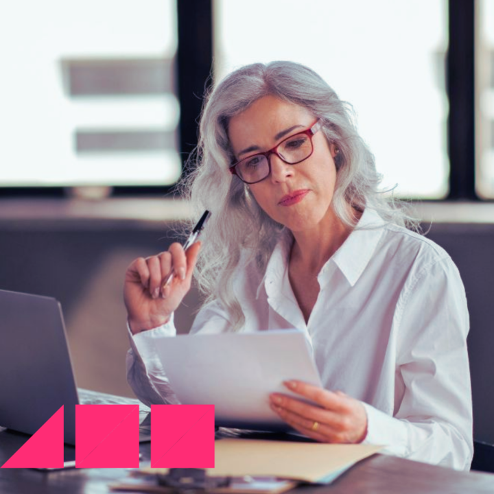 Woman wearing a white blouse and red glasses sitting at her desk with a pen in her hand and holding a physical copy of her return to workforce cover letter