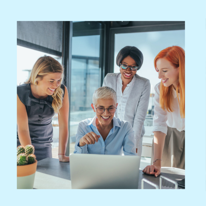 Three business woman in an office hovering over another professional during her returnship and showing her how to complete a task on her laptop