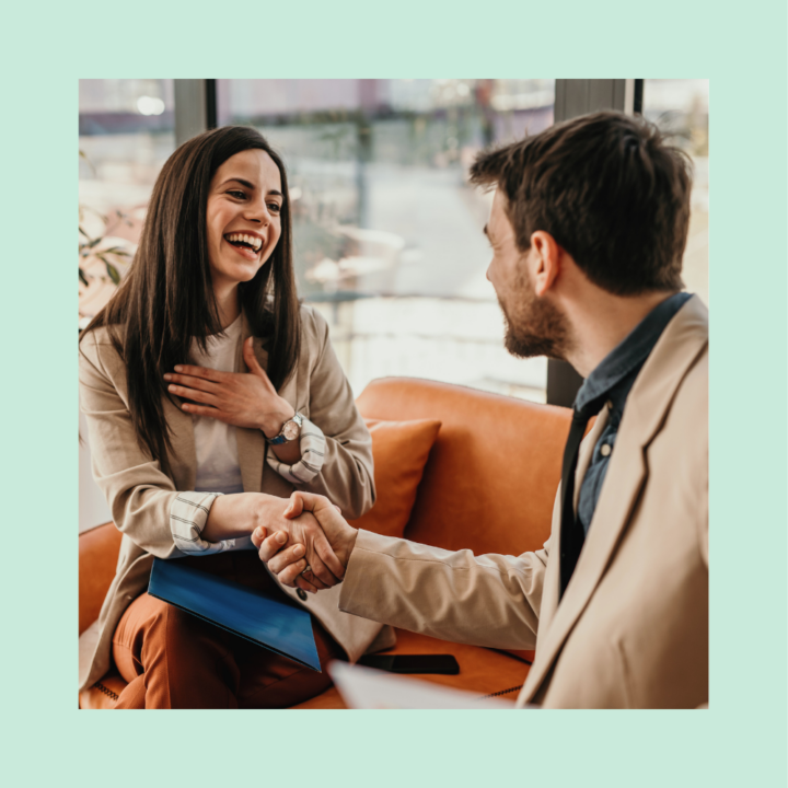 A smiling woman in professional attire shakes hands with a bearded man in a business setting. She holds a blue folder and appears grateful, while the man, also dressed in business attire, engages in friendly conversation. They are seated on an orange couch in a well-lit office with large windows in the background.