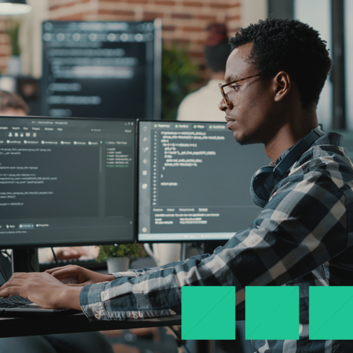 A full-stack developer wearing glasses and headphones works at a dual-monitor setup, coding in a modern office environment with a blurred colleague in the background.