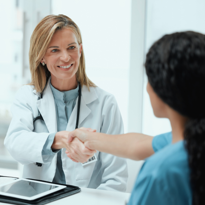 Smiling healthcare professional in a white coat shaking hands with a nurse during a job interview in a medical office.