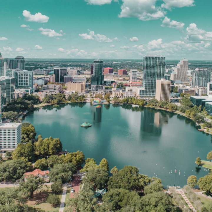 Aerial view of downtown Orlando, Florida, featuring high-rise office buildings surrounding Lake Eola with its iconic fountain and swan-shaped paddle boats. The scene is framed by trees, parks, and a bright blue sky with scattered clouds.
