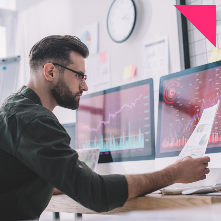 Male data analyst reviewing graphs on dual monitors while holding a printed report in a modern office setting