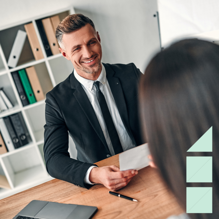 A professional man headhunter in a suit smiling and holding a resume during a job interview with a female candidate across the desk in a modern office setting.