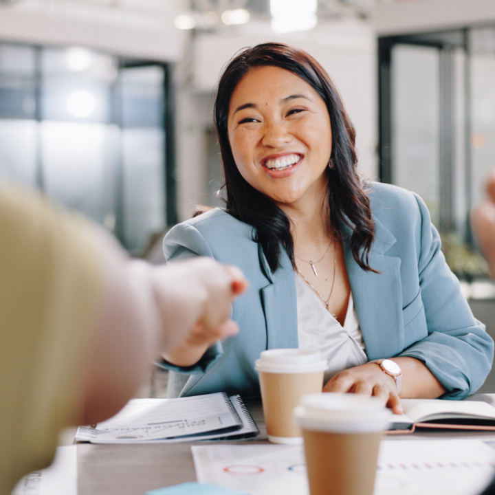 A woman in a blue blazer smiles and shakes hands with an interviewer across a desk with coffee cups and documents, illustrating a job interview during the process of recruiting HR professionals.