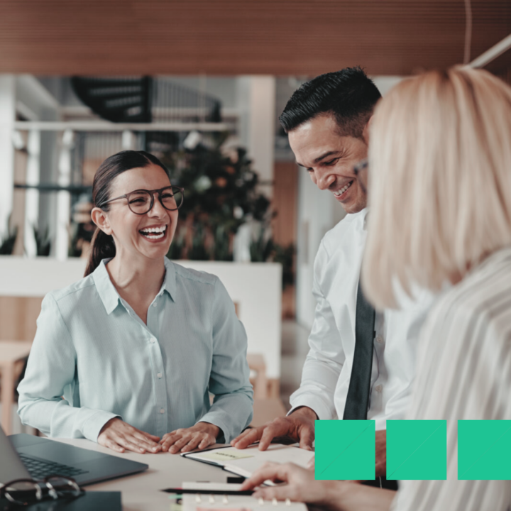 temporary and permanent employees around a desk in an office laughing and communicating about a project they are working on
