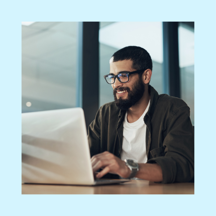 Smiling male hiring manager with glasses working on a laptop posting a role on an IT job board in a modern office setting, wearing a white t-shirt and dark jacket.