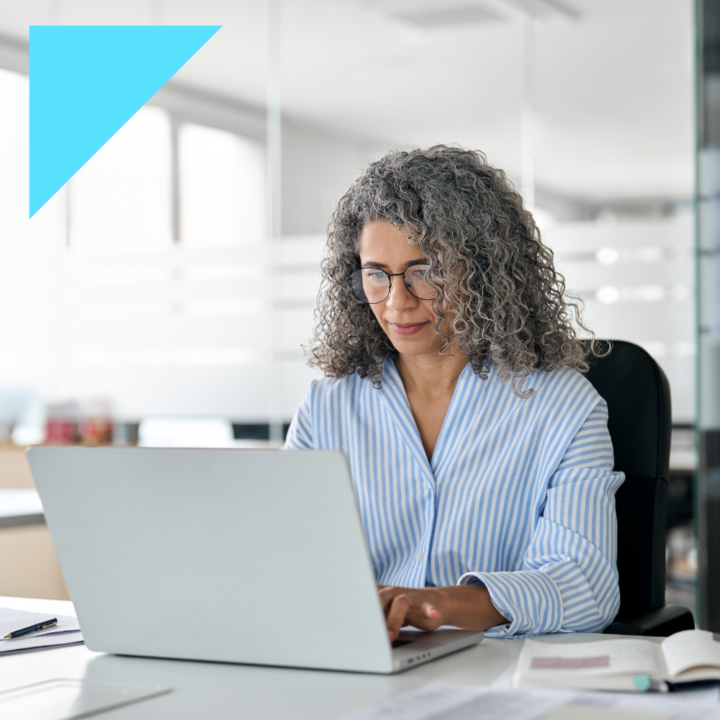 Professional hr manager woman with curly gray hair and glasses working on a laptop in a modern office. She is wearing a blue and white striped blouse and appears focused as she types a job description, with an open notebook and pen on the desk beside her.