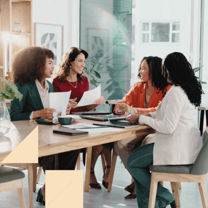Four professionally dressed recruiting women sit around a table in a bright, modern office space, engaged in a lively discussion. They are holding papers and notebooks, suggesting a collaborative meeting or interview. The atmosphere appears friendly and productive, with natural light streaming through large windows in the background.