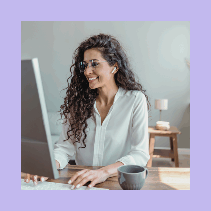 Smiling executive assistant woman with long curly hair wearing glasses and earbuds, working at a desktop computer in a bright home office. She is dressed in a white blouse and has a coffee mug on the desk in front of her.