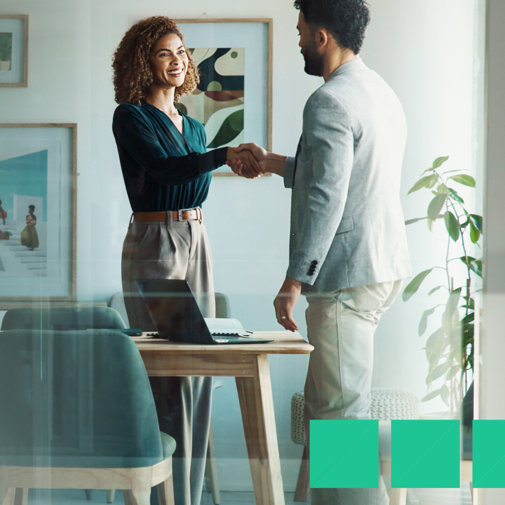 A woman and man smiling and shaking hands in a modern office setting, concluding a successful hr manager job interview. A laptop is open on the desk, with artwork and plants in the background.