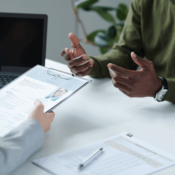 A close-up of a job interview in progress, showing a candidate gesturing while speaking across the table from an interviewer holding a clipboard with a printed CV. A laptop and documents are visible on the white desk, emphasizing a professional and evaluative setting.
