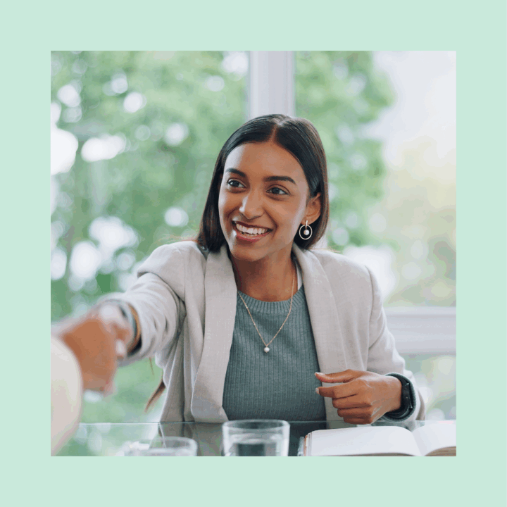 A smiling young recruiter woman in business attire reaches out to shake hands during a job interview. She is seated at a glass table with a notebook and water glass in front of her, with a bright window and greenery in the background.