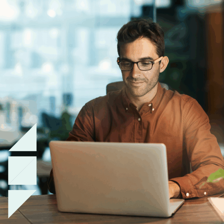 A professional recruiter man wearing glasses and a rust-colored shirt works on a laptop in a modern office, appearing focused and thoughtful, representing the challenge of finding qualified job candidates.