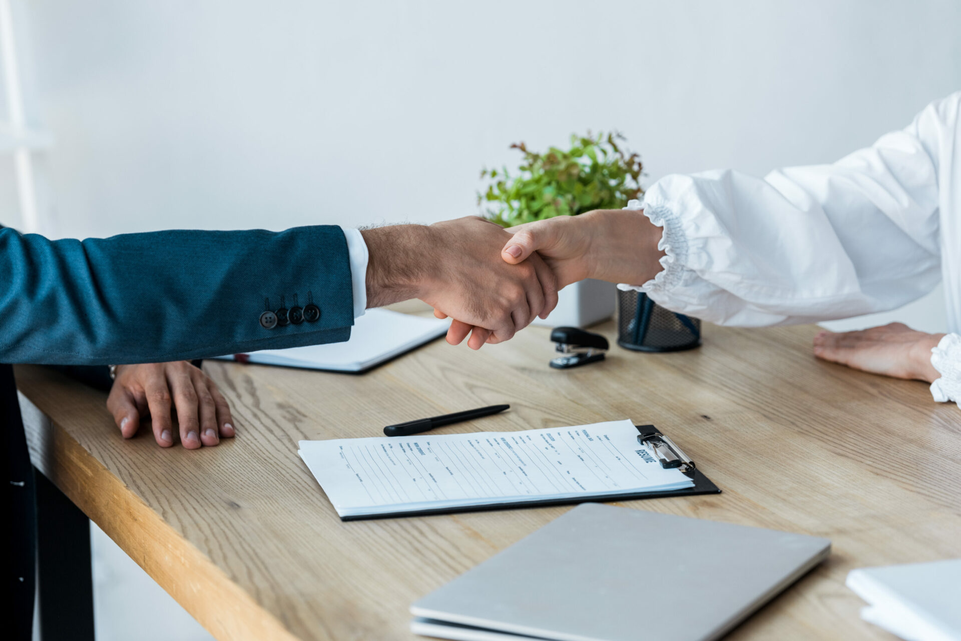 Close-up of two people shaking hands across a wooden desk, with a resume, pen, and laptop visible, symbolizing the conclusion of a successful job interview