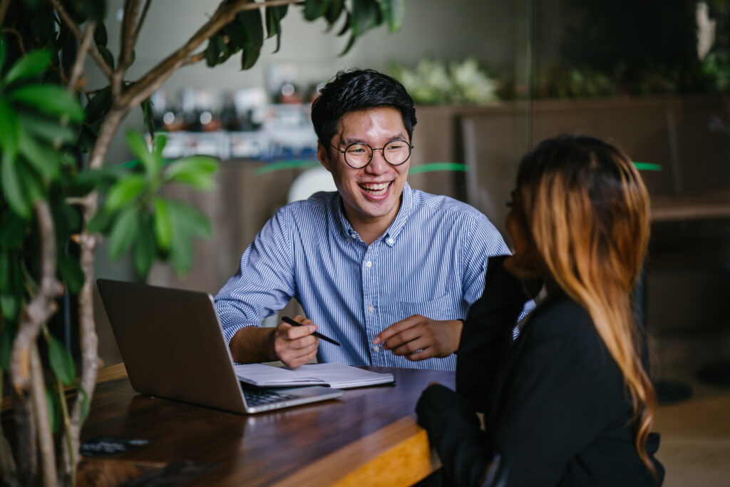 A RPO male recruiter in glasses and a striped shirt conducts an interview with a woman across the table, using a laptop and notebook, in a modern office space with plants and natural lighting.