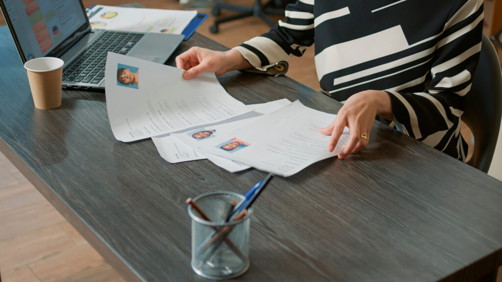 A recruiter reviews printed resumes with candidate headshots at a desk, next to an open laptop, coffee cup, and documents