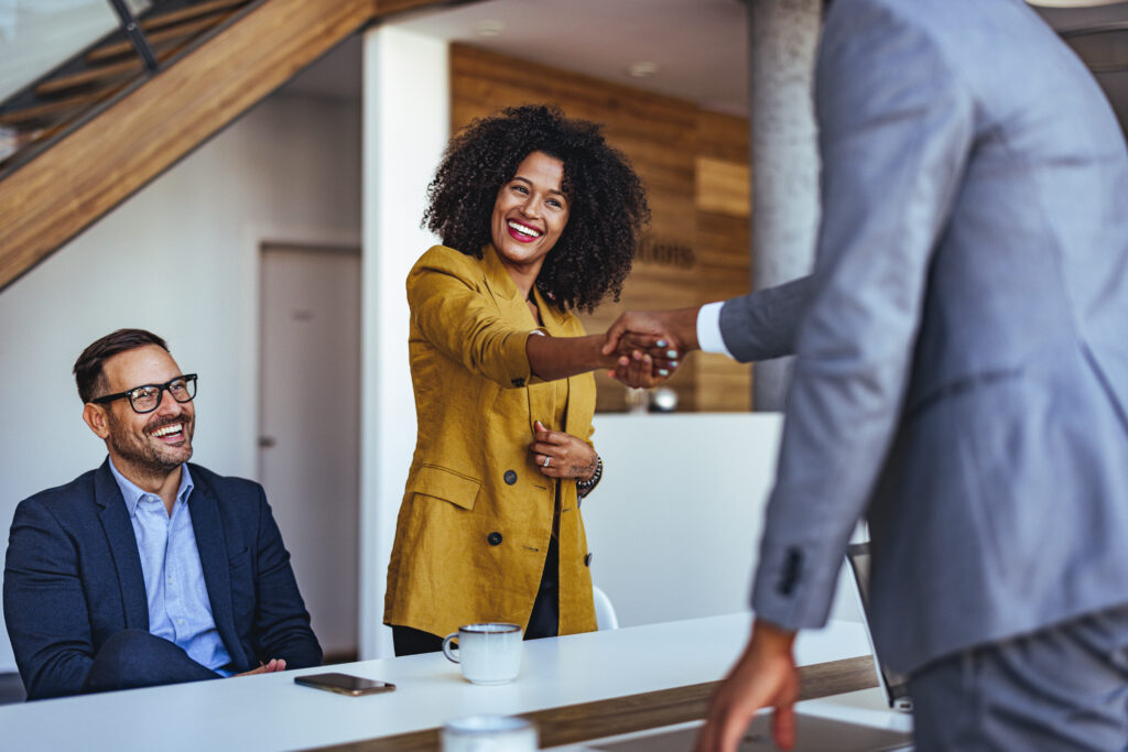 A professional woman in a mustard blazer smiles warmly as she shakes hands with a man in a suit, concluding a successful executive interview or meeting, while another man seated nearby watches and smiles in a modern, stylish office setting.