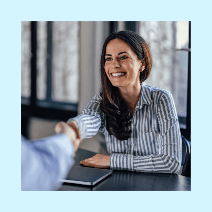 A smiling woman in a striped shirt shakes hands across a desk during a meeting, symbolizing a successful interview or onboarding process—representing the growing trend of fractional hiring to access top talent on a flexible basis.