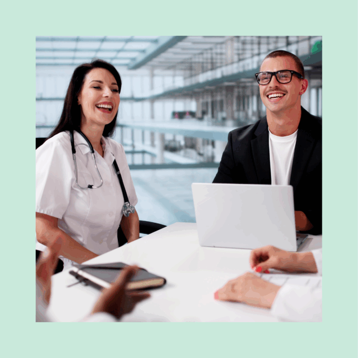 A group of staffing professionals, including a smiling female doctor with a stethoscope and a man in business attire with a laptop, sit around a table in a modern healthcare office setting, engaged in a job interview for healthcare admin role.