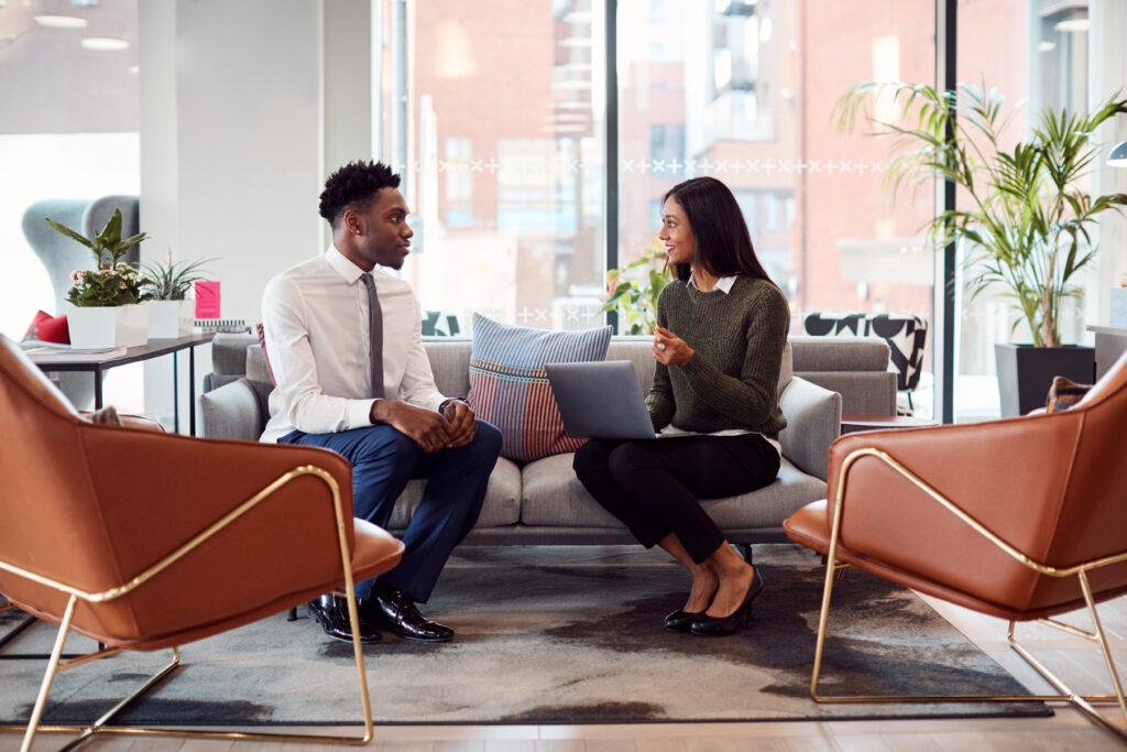A professional man and woman seated on a modern office couch engaged in a conversation, with the woman holding a laptop and gesturing as she speaks, suggesting a job interview or business discussion. The setting includes stylish furniture, indoor plants, and large windows with a city view.