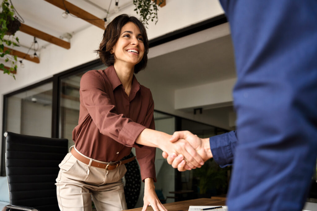 A smiling woman in business casual attire leans forward across a desk to shake hands with a man, indicating the start or end of a successful interview, in a modern office setting with natural light and greenery.