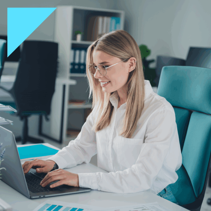 A smiling young professional woman wearing glasses and a white blouse works on a laptop at a modern office desk, with charts and documents visible in the foreground.