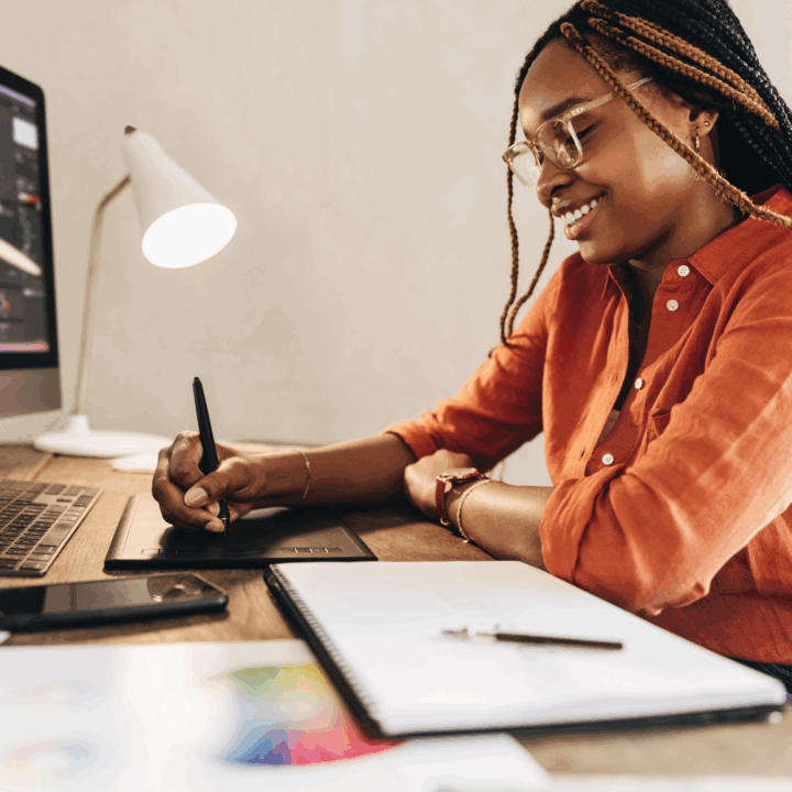 Smiling graphic designer using a digital drawing tablet at her desk, working on a branding project with a color wheel and sketchpad in view, ideal for illustrating creative work or hiring design talent.