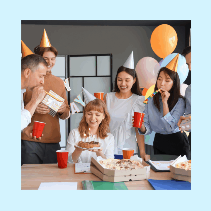 Group of coworkers wearing party hats celebrating a birthday in an office setting. A woman in the center is smiling and holding a small cake with lit candles, while others cheer with red party cups, balloons, and noisemakers. Pizza boxes and notebooks are on the table.