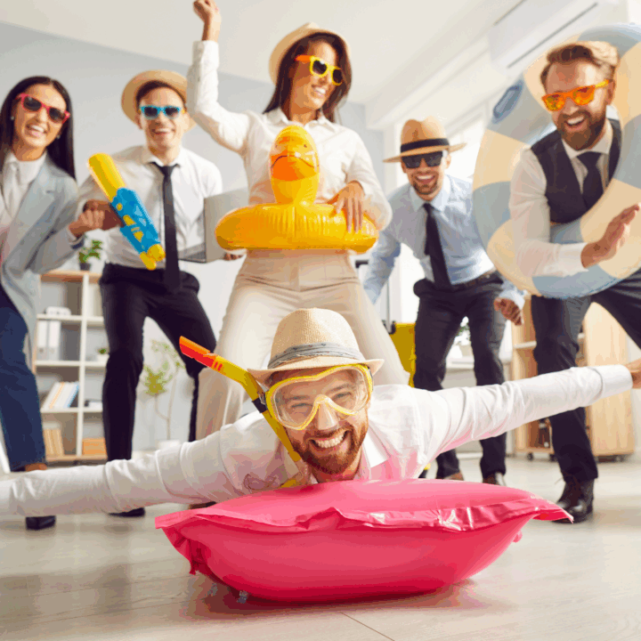Group of office employees dressed in business attire with summer accessories like goggles, floaties, and beach hats, playfully posing and laughing at a summer-themed work party indoors.