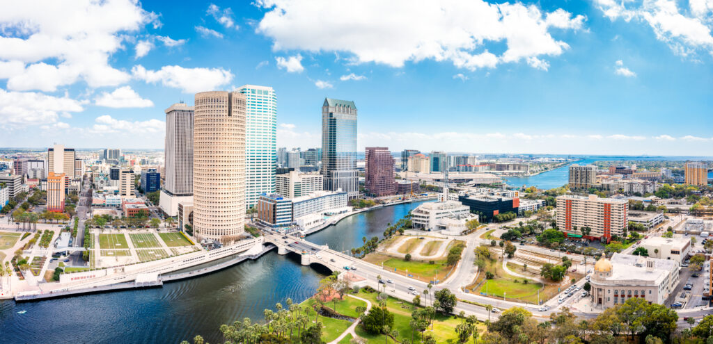 Panoramic view of downtown Tampa, Florida, featuring modern high-rise buildings, the Hillsborough River, bridges, and a bright blue sky with scattered clouds.