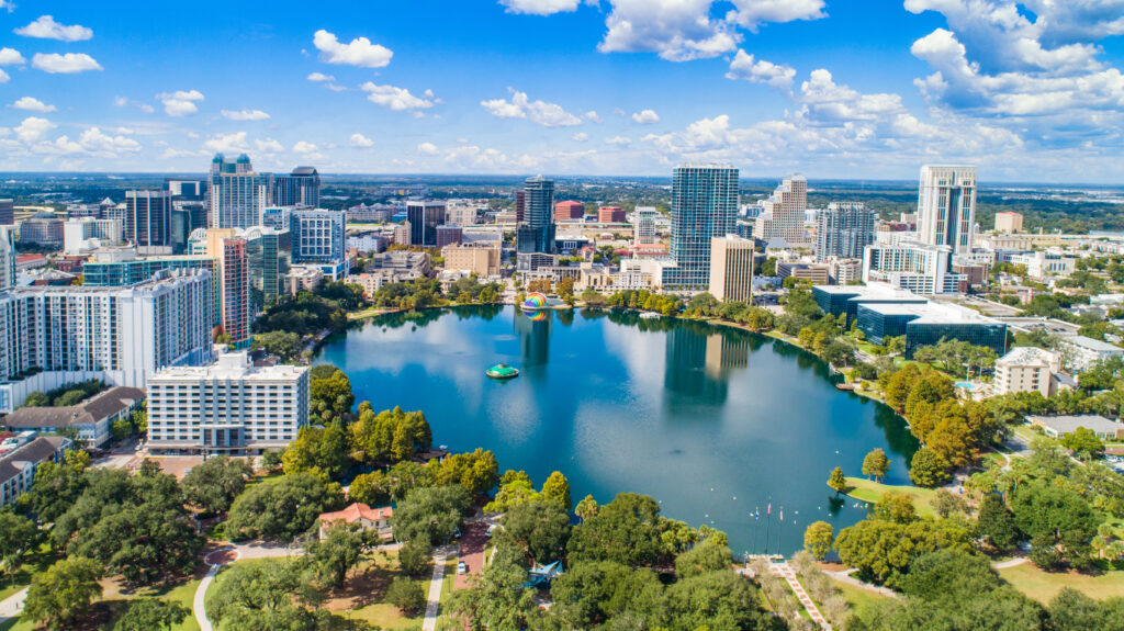 Aerial view of downtown Orlando, Florida, featuring Lake Eola with its iconic fountain and surrounding park, framed by modern high-rise buildings under a bright blue sky with scattered clouds.