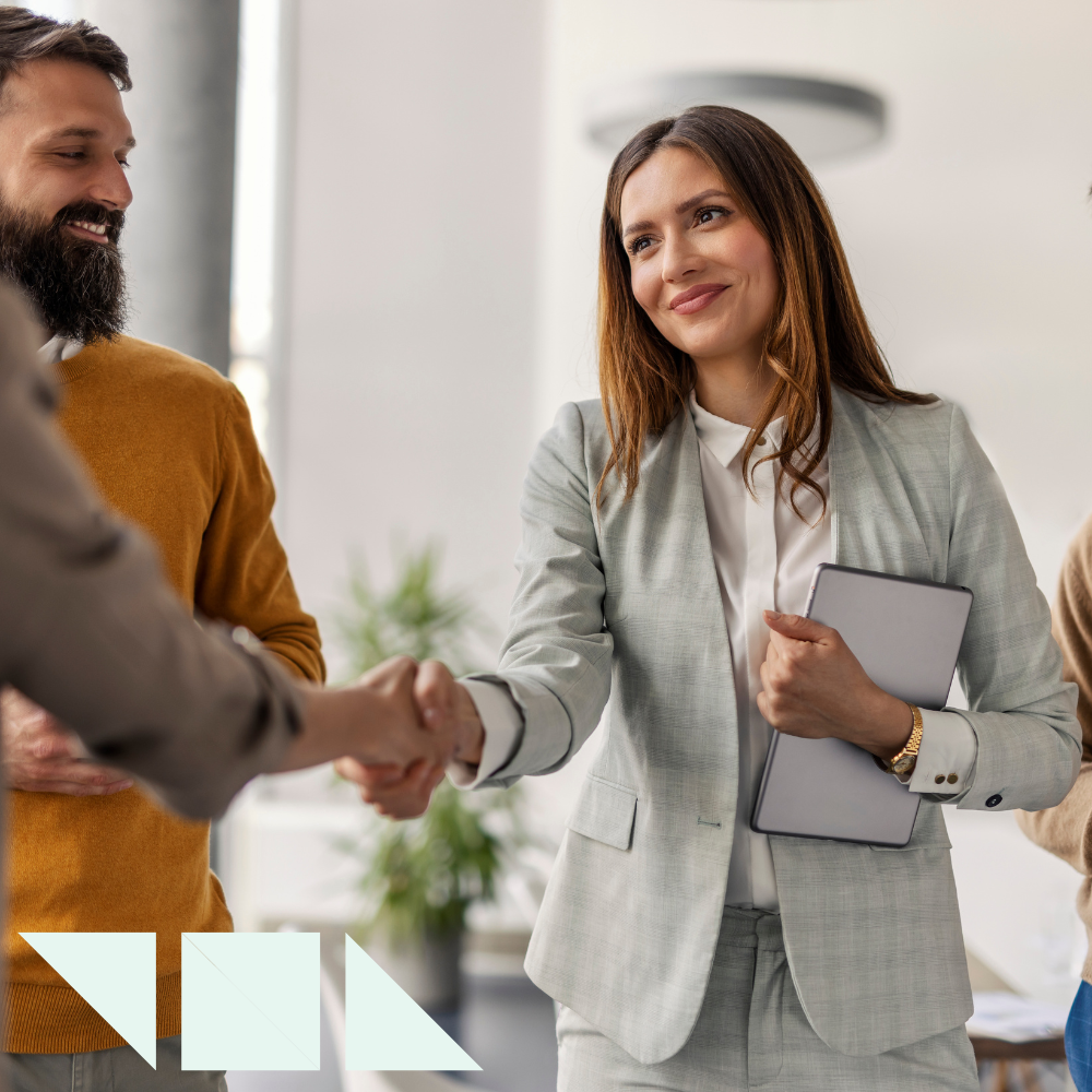 A professional hr recruiter woman in a light gray suit shakes hands with an hr professional during a business meeting, holding a tablet under her arm and smiling confidently