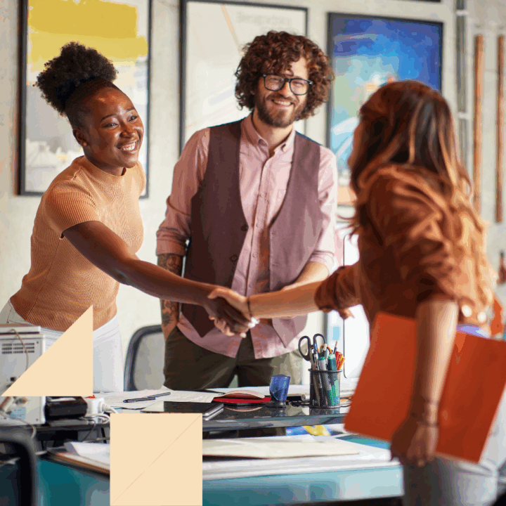 A woman shakes hands with a job candidate during a marketing team interview in a creative office setting, while a team member looks on and smiles in the background. The atmosphere appears friendly and professional.