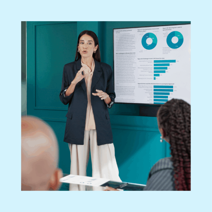 Female operations manager professional giving a presentation with data charts displayed on a screen, speaking to colleagues in a meeting room.