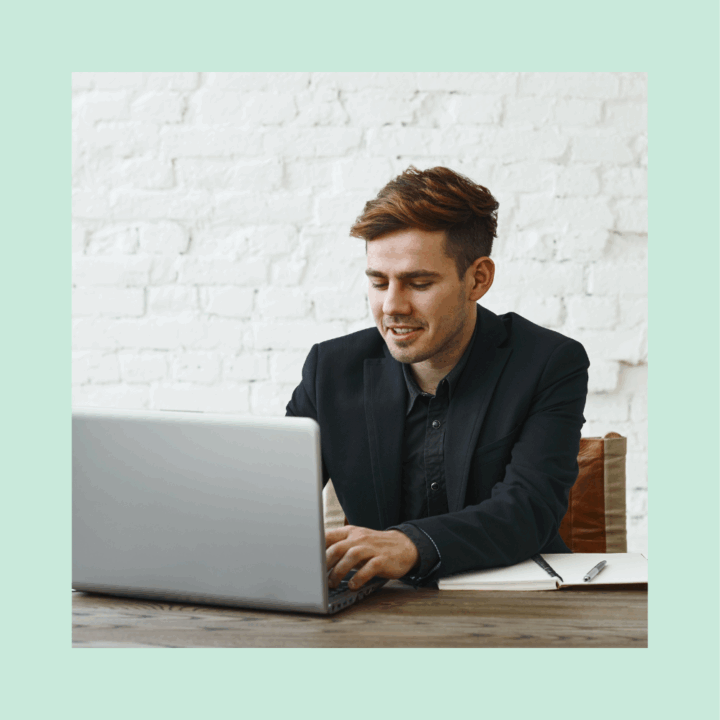 Smiling man in a black suit sitting at a wooden desk, typing on a laptop with a notebook and pen beside him. He is seated in front of a white brick wall, appearing engaged and focused on using machine learning for his recruiting efforts