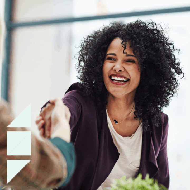 A smiling woman with curly hair reaches out to shake hands with another person during what appears to be a professional meeting or job interview. She is wearing a dark blazer and a light-colored top, and the atmosphere is bright and friendly with natural light in the background.