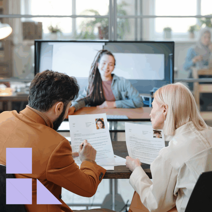 Two hiring managers sit at a table reviewing resumes while conducting a video interview with a candidate on screen, focusing on her education background and academic qualifications as part of the hiring discussion.