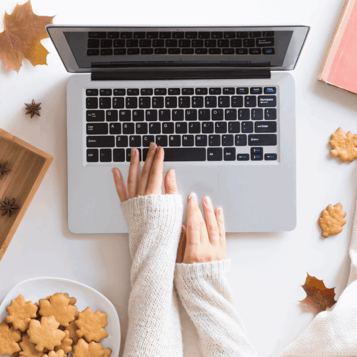 Top view of a person typing on a laptop with cozy white sweater sleeves, surrounded by autumn-themed cookies, star anise, and fall leaves on a white desk.