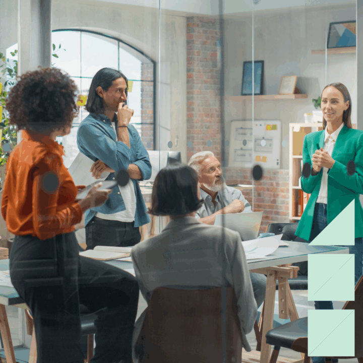 A diverse marketing team meets in a modern office conference room. A woman in a green blazer leads the discussion while colleagues listen, take notes, and collaborate on strategy around a shared table.