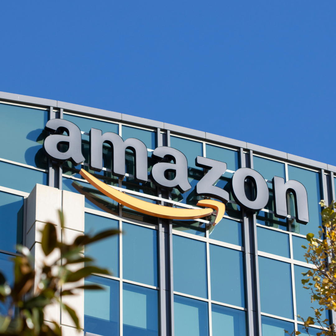 The Amazon logo is displayed on the exterior of a modern glass office building, with blue sky overhead and trees partially visible in the foreground.
