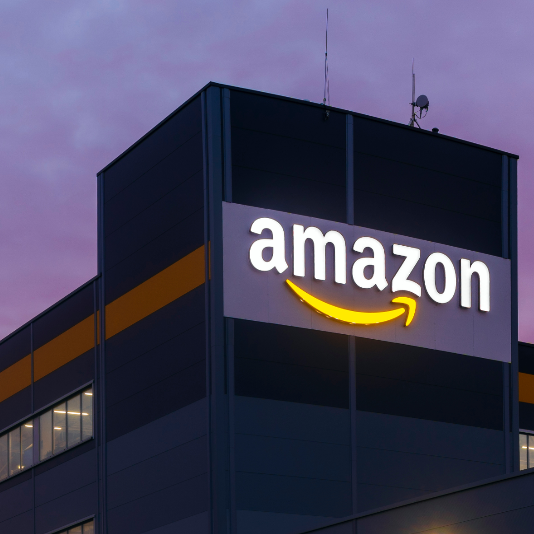 An Amazon office or warehouse building is shown at dusk, with the illuminated Amazon logo mounted on the exterior against a purple evening sky.