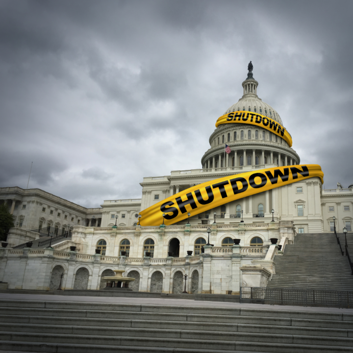 The U.S. Capitol building is shown under a cloudy sky, with large yellow banners reading “SHUTDOWN” wrapped across the dome and façade, symbolizing a government shutdown.